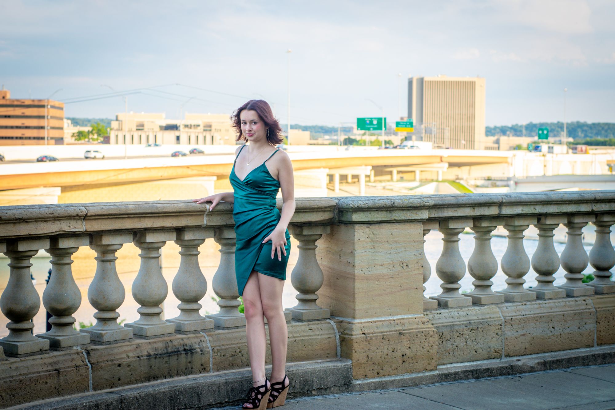 A high-school senior in a green dress leaning against a stone balustrade with the Dayton skyline and river behind her