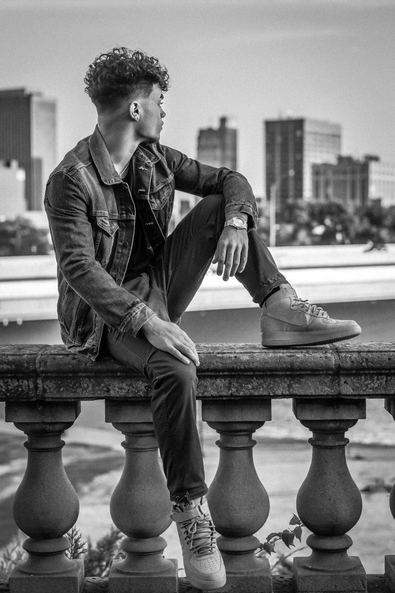 Black and white photograph of a senior seated on the stone balustrade above the river, city buildings behind him