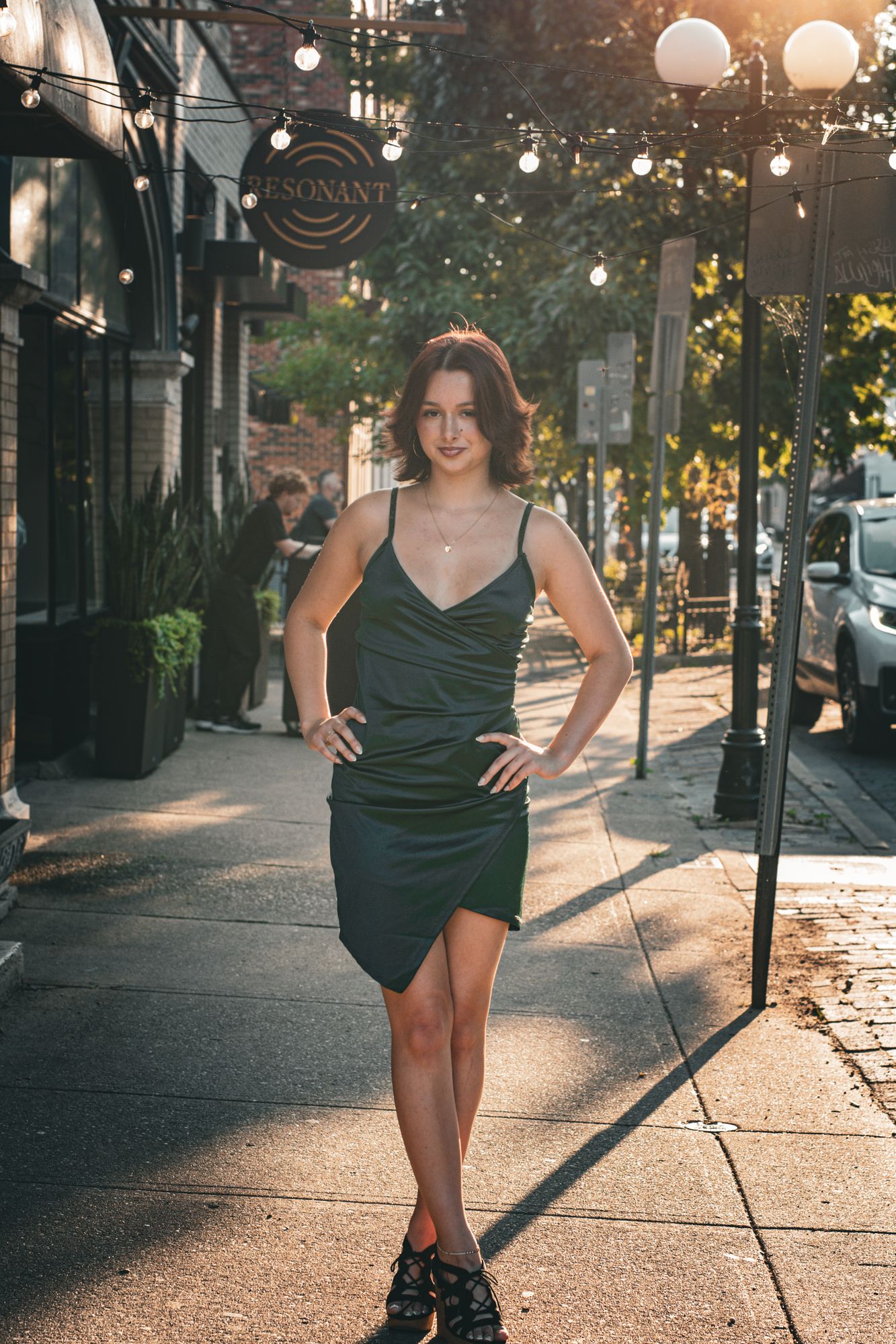A high-school senior standing on the sidewalk under string lights outside a brick storefront in low evening sun