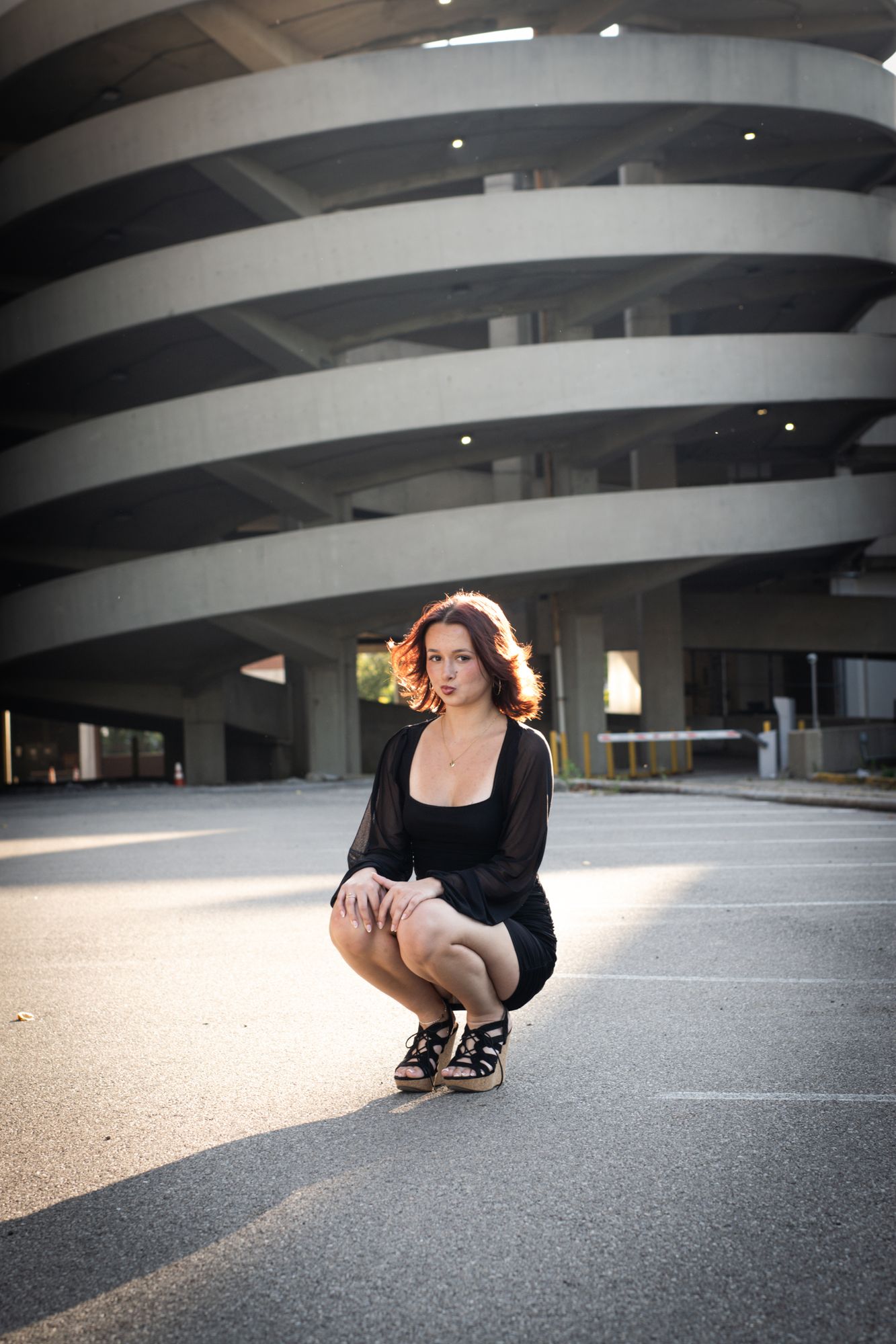 A senior crouched in an empty lot with the sweeping concrete curves of a parking structure rising behind her