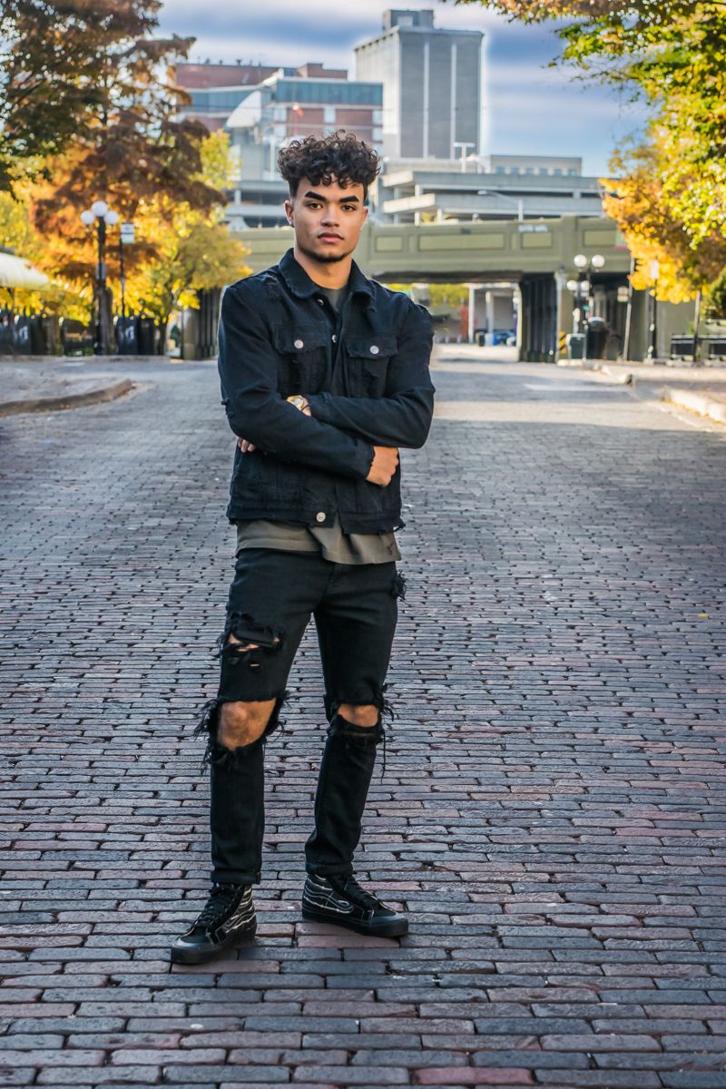 A high-school senior standing in the middle of a brick-paved street, autumn trees and the downtown skyline behind him