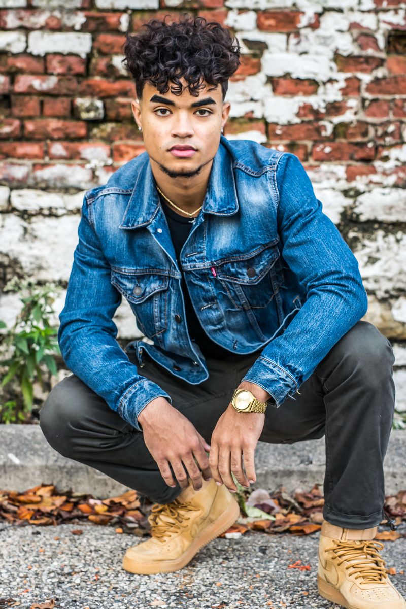 A senior in a denim jacket crouched against a weathered red and white painted brick wall