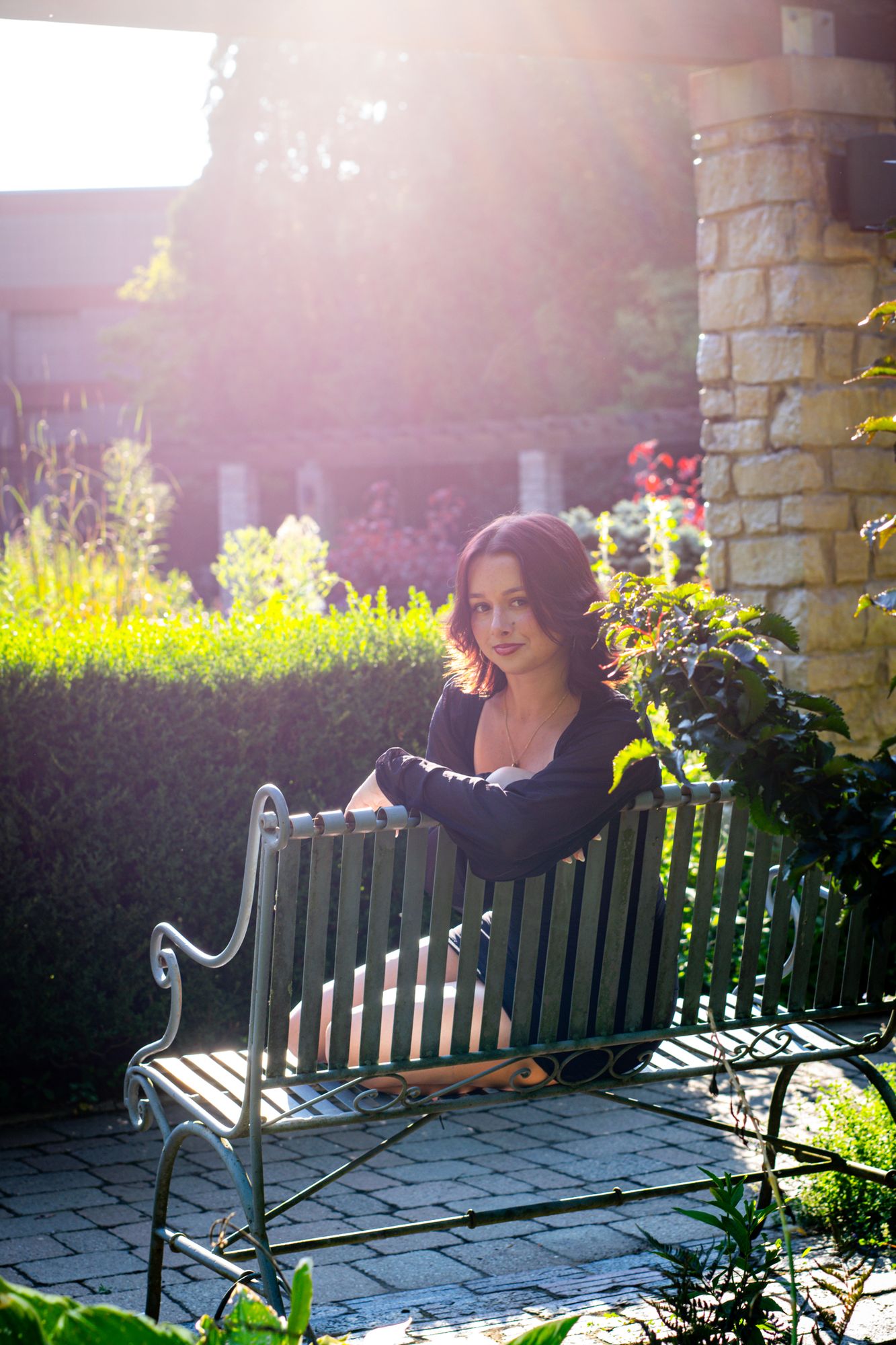 A high-school senior seated on an ornate iron bench in a garden, backlit by low evening sun