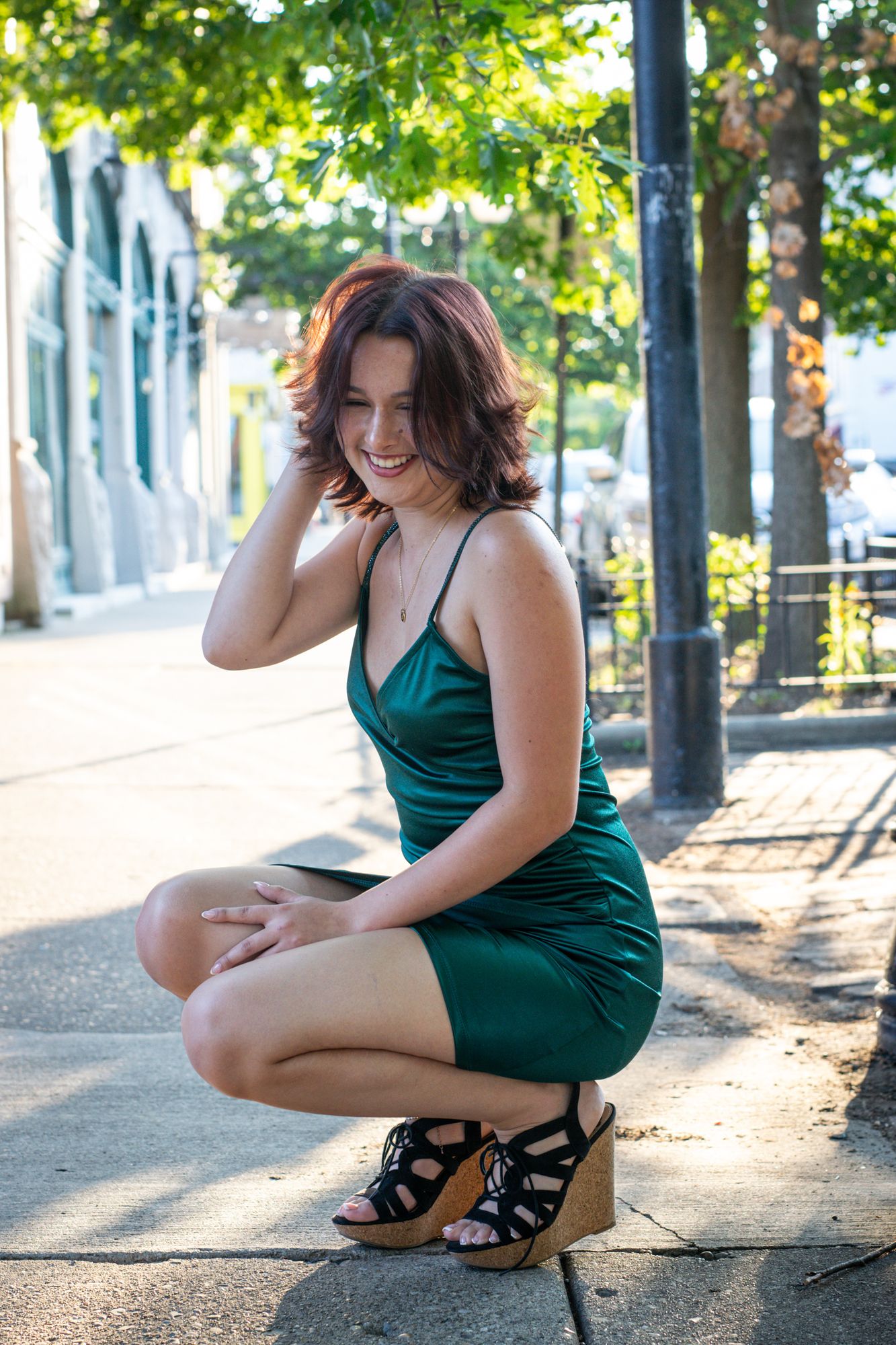A high-school senior crouched on a city sidewalk, laughing, backlit by late afternoon light