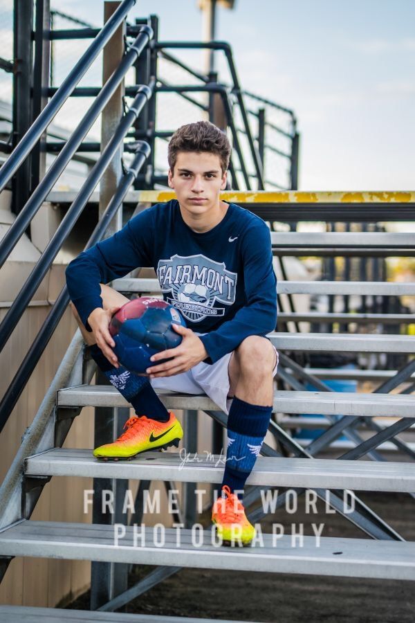 A high-school senior in a soccer jersey and cleats seated on stadium bleachers, holding a ball