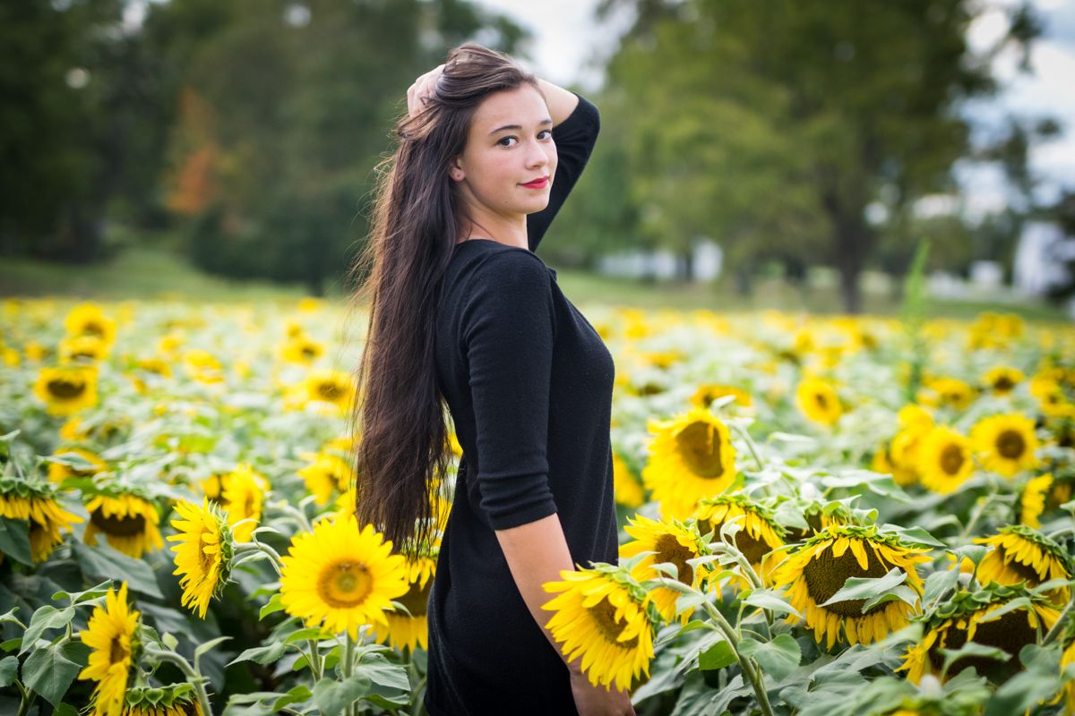 A high-school senior in a black dress turning back toward the camera in a field of sunflowers