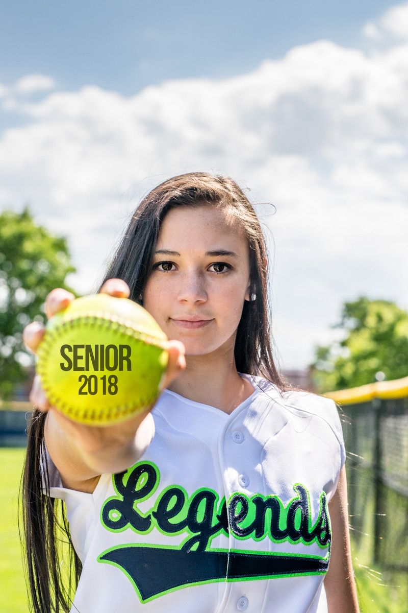 A high-school senior in a softball jersey holding a ball out toward the camera on a sunlit field